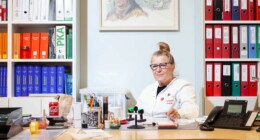 Auf dem Bild sitzt die Apothekerin an ihrem Tisch. Hinter ihr ist ein grosses Regal